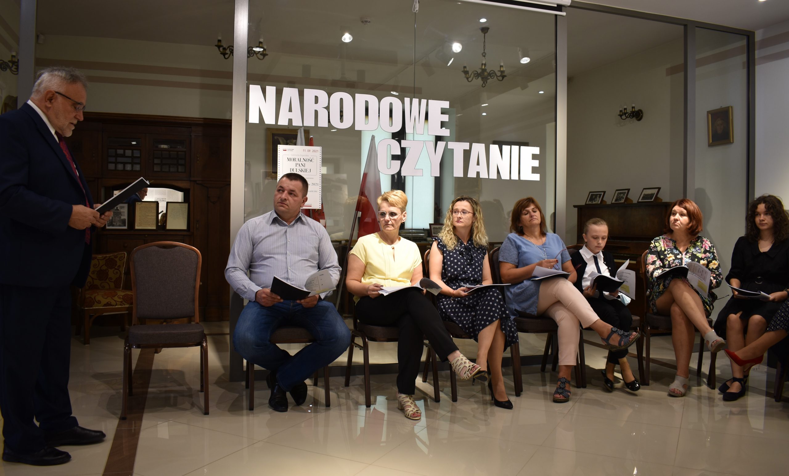 Pan Wójt czyta, reszta uczestników siedzi z zaciekawieniem słuchając. W tle eksponaty Muzeum Konstantego Laszczki