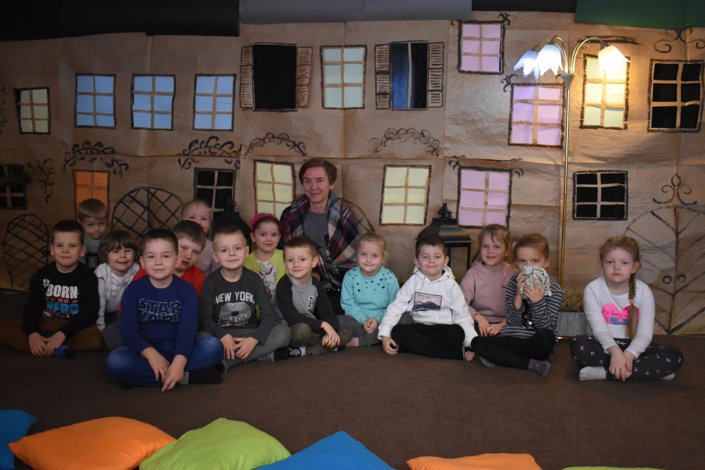 Na pierwszym planie dzieci siedzące na kolorowych poduszkach, w tle dekoracja przedstawiająca kamienice, rozświetlone lampiony i postać w przebraniu z bajki.