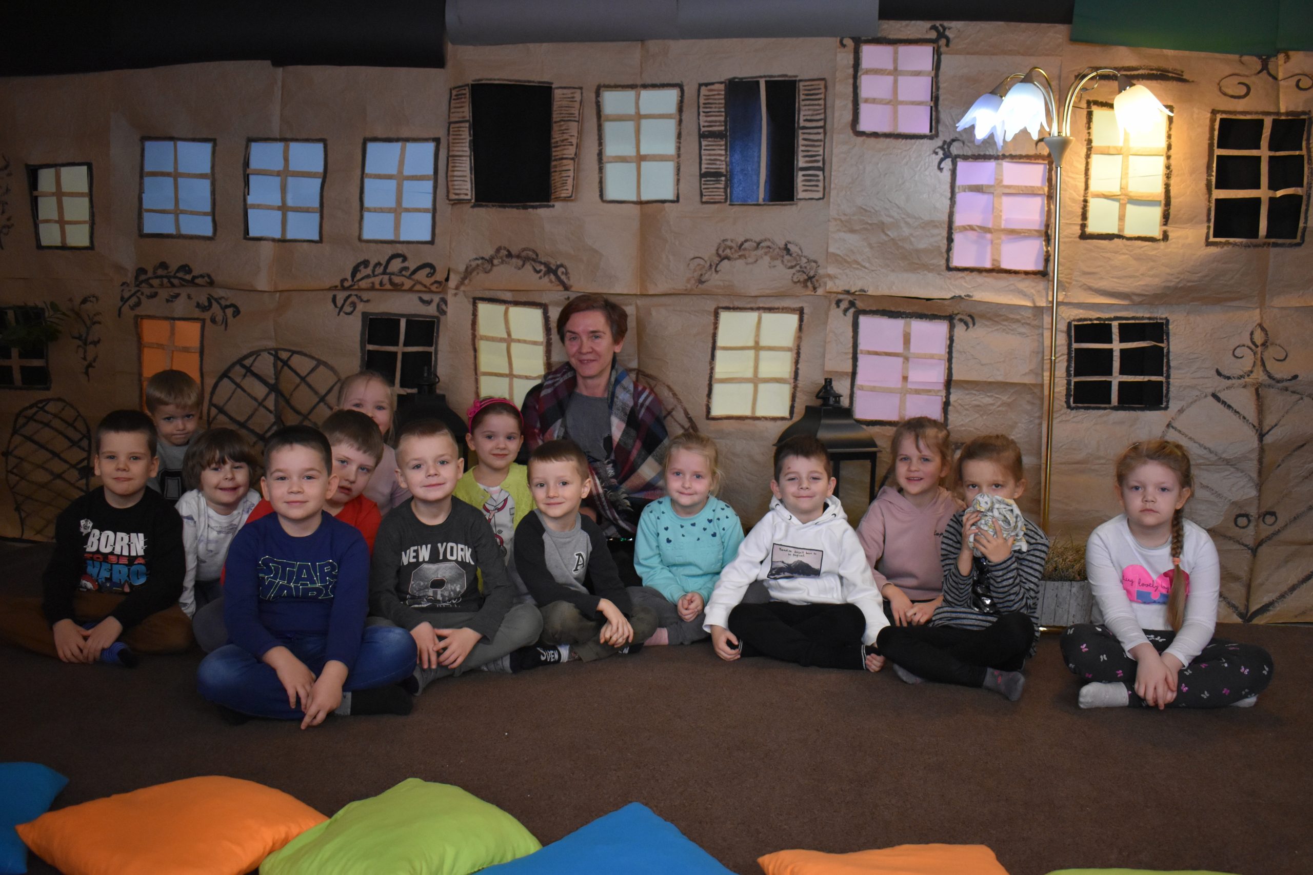 Na pierwszym planie dzieci siedzące na kolorowych poduszkach, w tle dekoracja przedstawiająca kamienice, rozświetlone lampiony i postać w przebraniu z bajki.