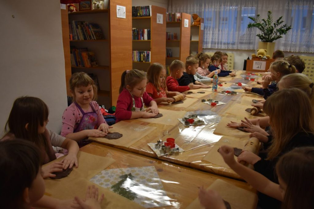 Stroiki świąteczne z gałązek świerkowych, bombek, orzechów, pomarańczy na drewnianym krążku ze świeczką. Na pierwszym planie stoi choinka z piernika zapakowana w folię z pomarańczową wstążką.