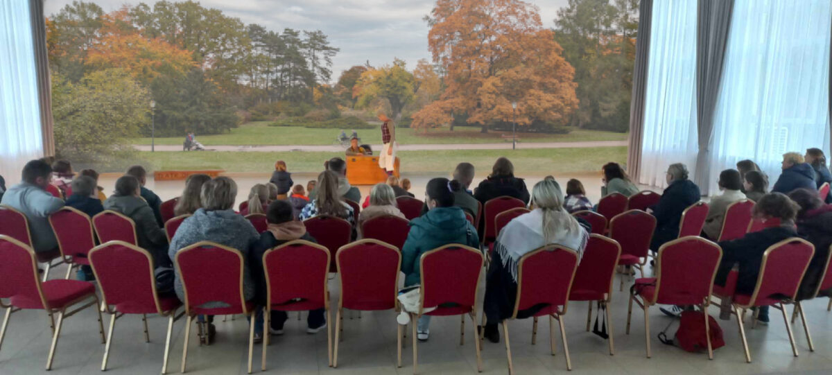 W sali na czerwonych krzesłach siedzi siedzą osoby i oglądają występ dwóch mężczyzn. W tle obraz z jesiennym parkiem.