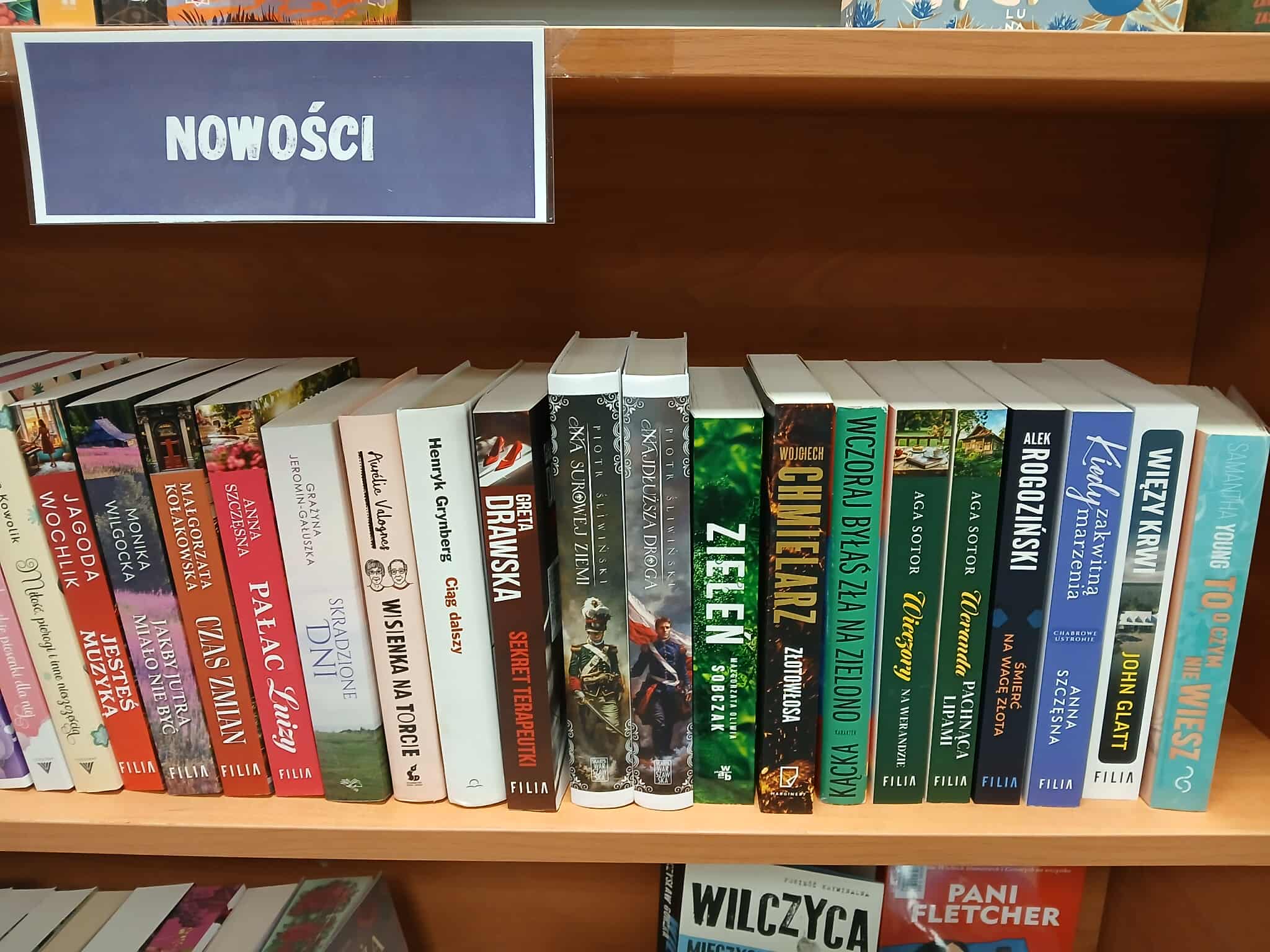 Półka z nowościami w księgarni, pełna kolorowych grzbietów książek. Tło to drewniana półka. Widoczny napis NOWOŚCI.