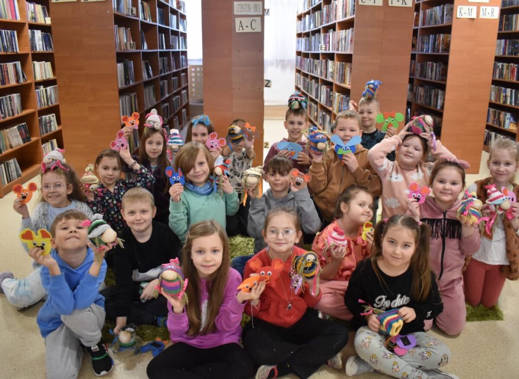 Uśmiechnięte dzieci pozują do zdjęcia z zrobionymi przez siebie skrzatami i magnesami. W tle biblioteczne regały z książkami.