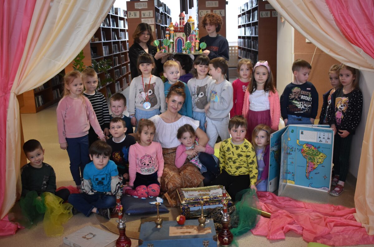 Gróbka przedszkolaków z kobietą w białej bluzce siedzą na dywanie reszta stoi. Przed nimi leżą kuferki czerwone wieże, duży atlas z mapą ameryki południową. Po obu stronach kolorowe kotary