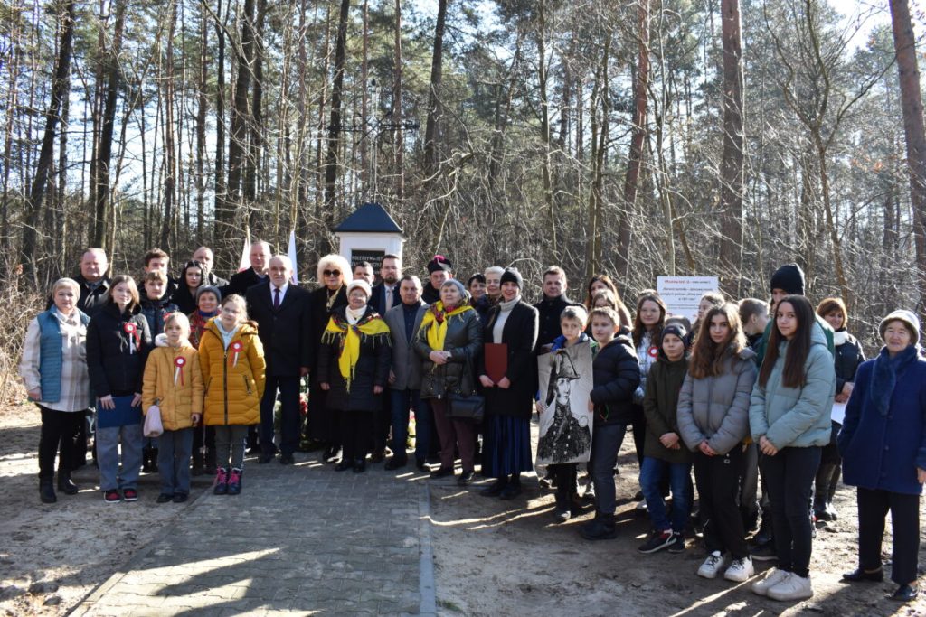 Na zdjęciu widać grupę ludzi, która stoi na terenie zalesionym. Przyrodniczy krajobraz powodujący zagrożenie dla ich spotkań lub spaceru.