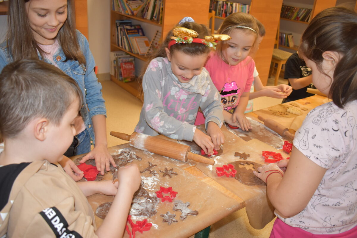 Dzieci w Bibliotece na drewnianym stole zabierają się do pieczenia pierników. W tle regały z książkami