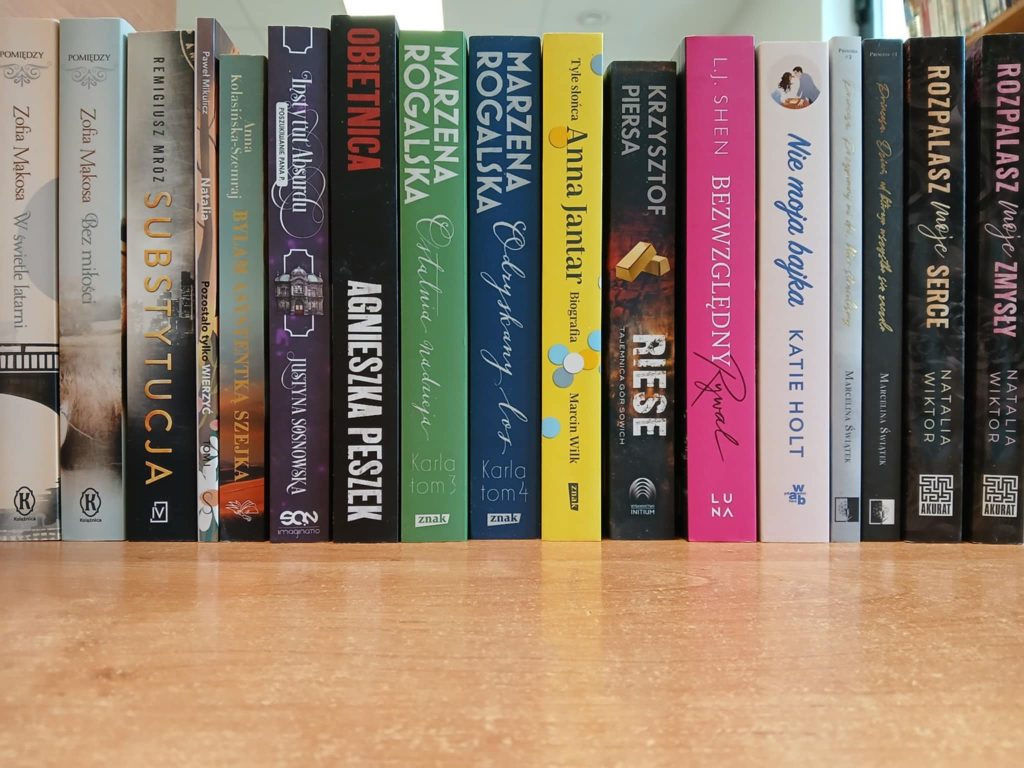 Rząd książek na półce z różnorodnymi okładkami. Kolory dominujące to czarny, zielony, biały i różowy. Tło to drewniana powierzchnia.