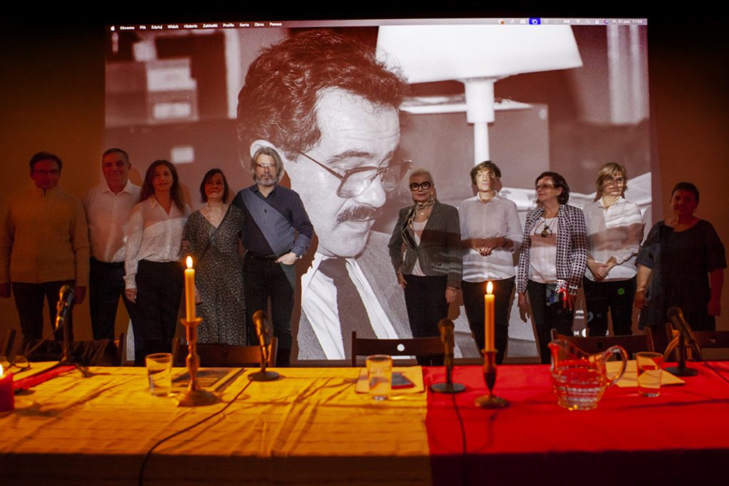 Grupa ludzi stoi przed dużą projekcją czarno-białego zdjęcia mężczyzny. Na pierwszym planie widoczne są oświetlone świecami stoły z mikrofonami. Tło jest ciemne, a projekcja dominuje w centralnej części obrazu.