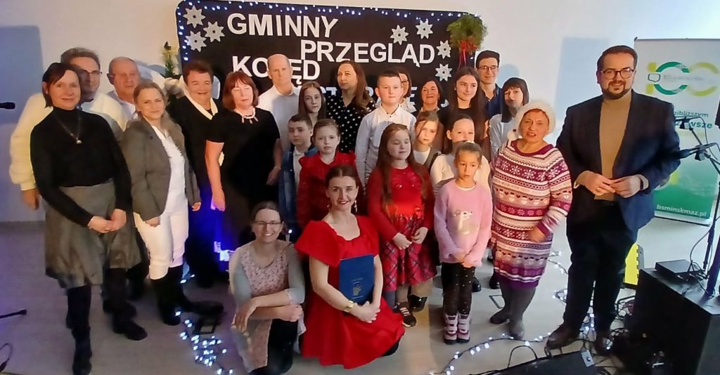 Na zdjęciu grupa osób na tle czarnego banera z napisem Gminny Przegląd. Ubrania w jasnych kolorach, w tym czerwona sukienka. Tło ozdobione światełkami i śnieżynkami.