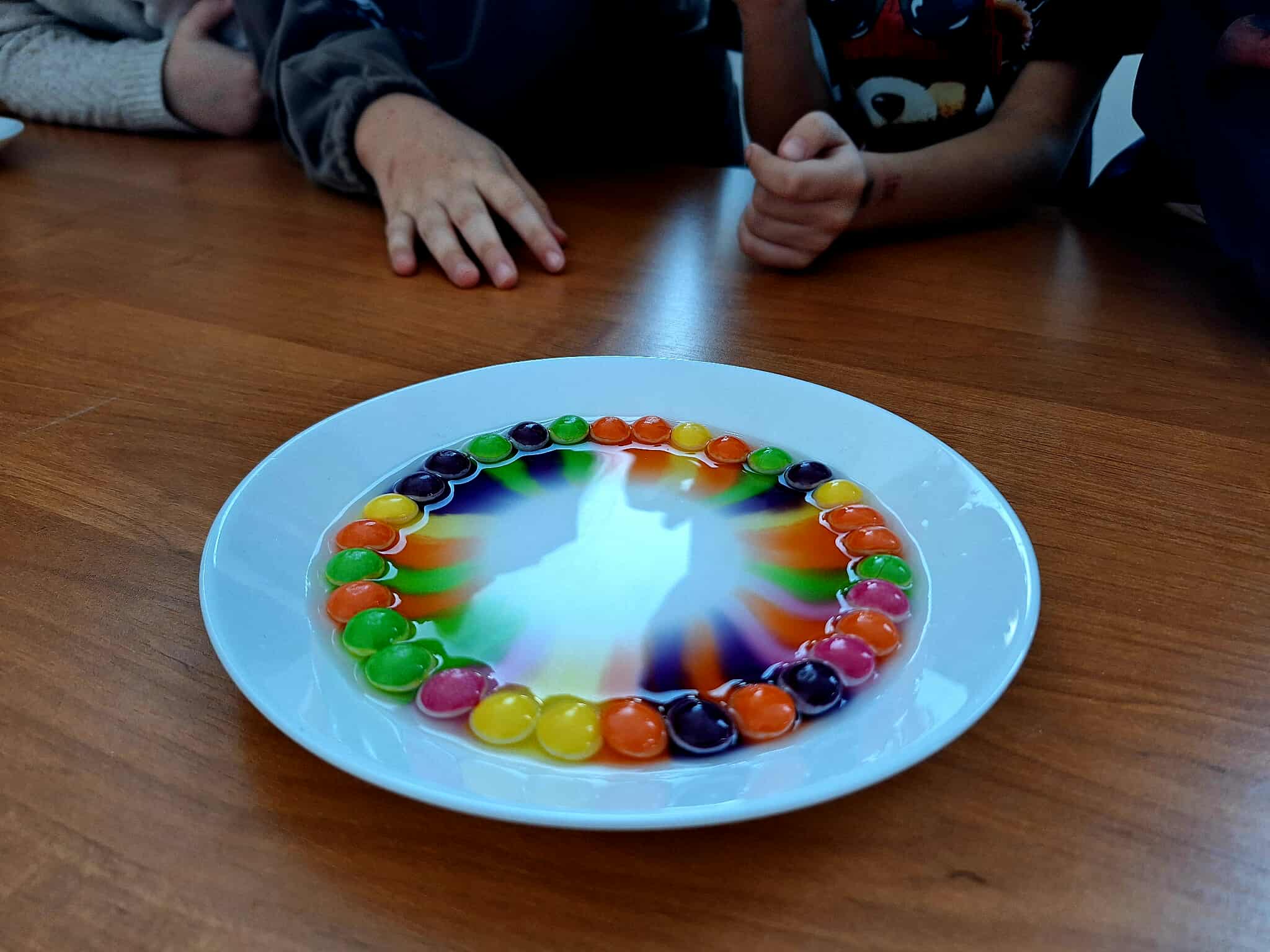 Biały talerz z m&msami w okręgu, tworzącymi tęczowy wzór w mleku. Drewniany stół, dłonie widoczne w tle. Kolory: różowy, fioletowy, zielony, pomarańczowy.