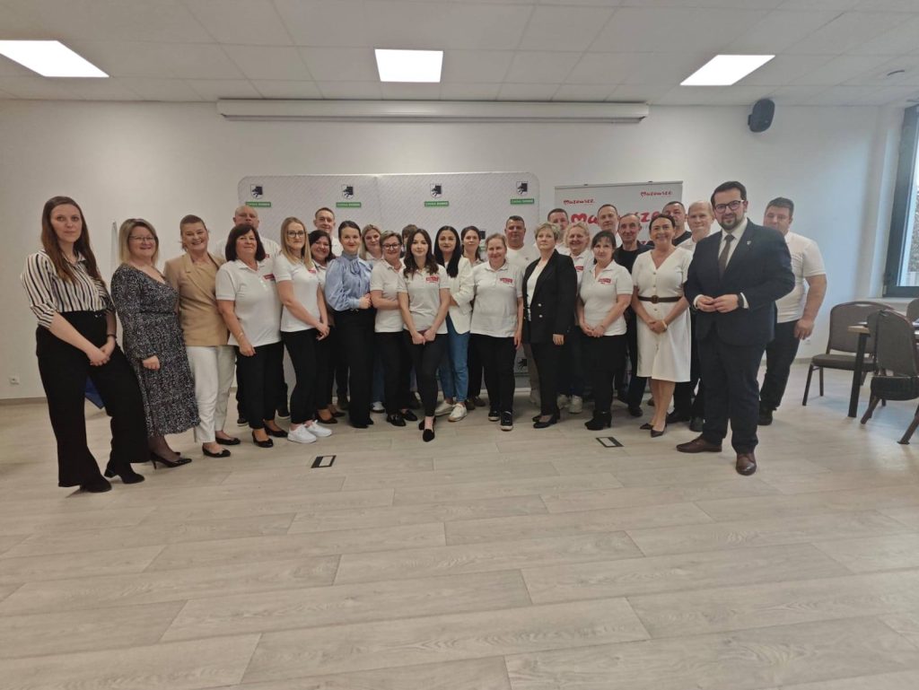 Grupa ludzi pozuje do zdjęcia w jasnym pomieszczeniu z drewnianą podłogą. Większość osób ubrana jest w białe koszulki. W tle widoczna jest szara ściana z logo.