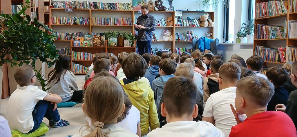 Grupa dzieci siedzi na podłodze w bibliotece z książkami na półkach. Dorosły mężczyzna czyta na tle pełnym kolorowych książek. Wokół rośliny i jasne światło z okna.