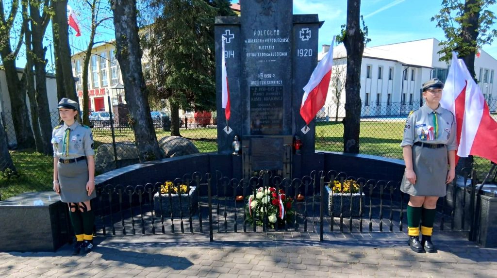 Pomnik z tablicą upamiętniającą, otoczony jest flagami Polski i kwiatami. Dwie harcerki w mundurach stoją na baczność. Tło stanowi ogrodzenie, drzewa i biały budynek.