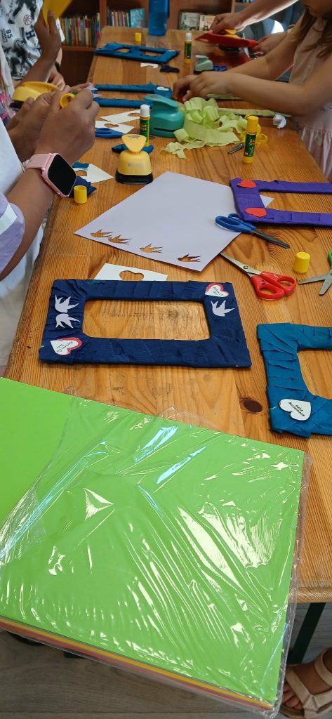 Kolorowe warsztaty plastyczne na drewnianym stole. Dzieci ozdabiają ramki wycinankami i klejem. Na stole zielony papier, nożyczki, klej.
