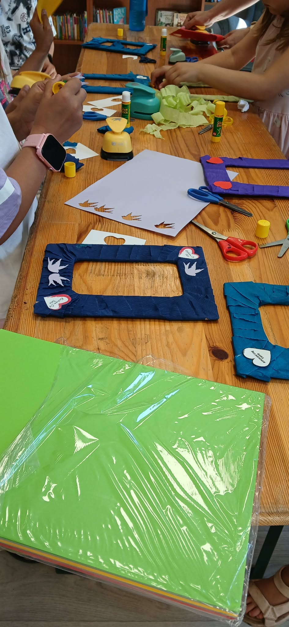 Kolorowe warsztaty plastyczne na drewnianym stole. Dzieci ozdabiają ramki wycinankami i klejem. Na stole zielony papier, nożyczki, klej.