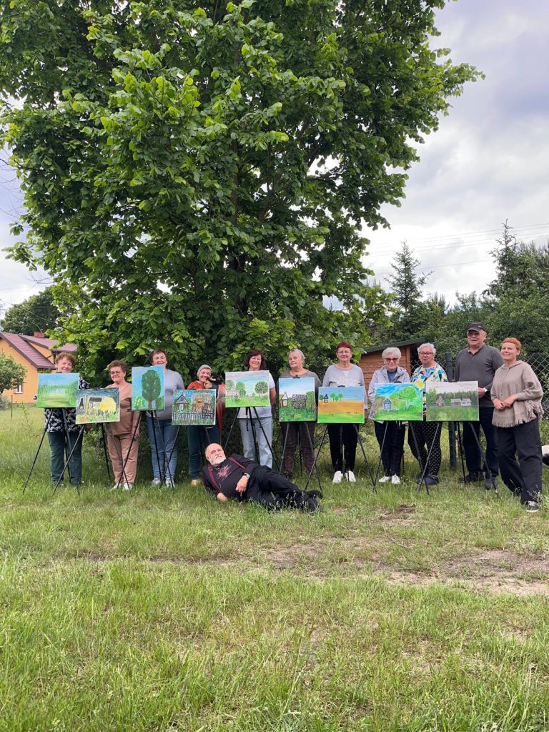 Grupa ludzi stoi na trawie przed drzewem z obrazami na sztalugach. Obrazy są kolorowe, dominują na nich zielone krajobrazy. W tle widać chmury i domki.