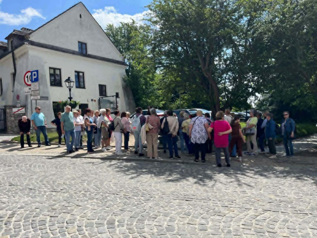 Grupa ludzi stoi na ulicy obok białego budynku. W tle drzewa i zaparkowane samochody. Na niebie widać chmury i słońce.