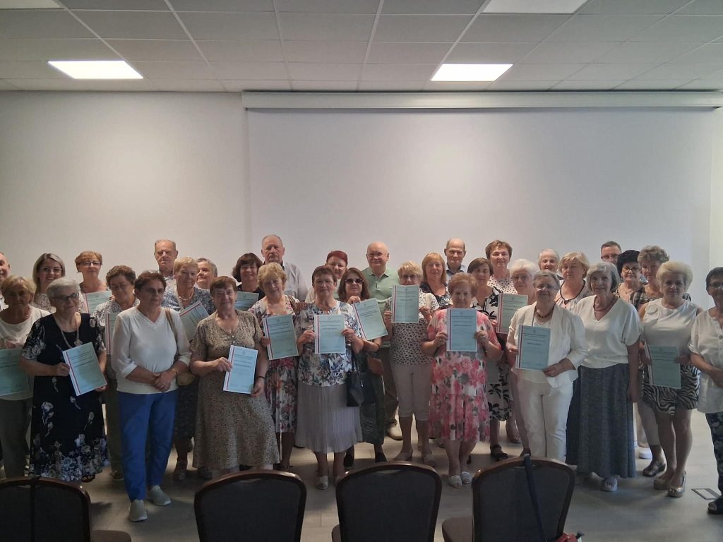Na obrazie grupa starszych osób trzyma dyplomy, stojąc przed białą ścianą w jasnym pomieszczeniu. Ubrania w stonowanych kolorach.