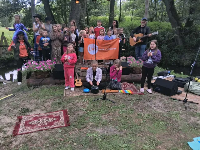 Grupa dzieci z instrumentami, w parku z zielonym tłem, przy pomarańczowym banerze. Kolorowe stroje i instrumenty. Akustyczna gitara i śpiew znakują wydarzenie.