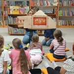 Dzieci siedzą na poduszkach w bibliotece, patrząc na osobę prezentującą teatrzyk kamishibai. Tło wypełniają kolorowe książki na półkach. Atmosfera jest przyjazna, a kolory są ciepłe i zachęcające.