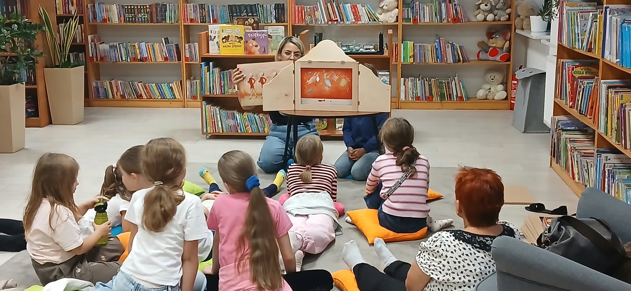 Dzieci siedzą na poduszkach w bibliotece, patrząc na osobę prezentującą teatrzyk kamishibai. Tło wypełniają kolorowe książki na półkach. Atmosfera jest przyjazna, a kolory są ciepłe i zachęcające.