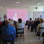Grupa ludzi siedzi w sali konferencyjnej, patrząc na prezentację z różowym slajdem. Kobieta prowadząca występuje przed ekranem. Tło jest jasne z oknami po prawej stronie.
