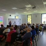 Pokój z ludźmi siedzącymi na krzesłach, patrzącymi na prezentację. Na ścianie wyświetlony jest kolorowy slajd. Przez okna widać zielone drzewa.