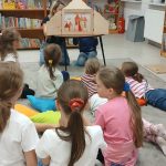 Dzieci siedzą na dywanie w bibliotece, oglądają teatrzyk. Kolorowe poduchy, półki z książkami w tle. Jasne wnętrze, dużo książek.