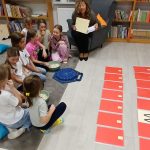 Grupa dzieci siedzi na dywanie z kolorowymi poduszkami, słuchając nauczycielki. W tle regał z książkami. Na podłodze układanki z czerwonych kart.