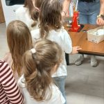 Osoby stoją w kolejce do stołu z urządzeniem. Tło to półki z książkami. Dominują kolory naturalne i czerwony.