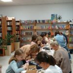 W pomieszczeniu z białym sufitem i regałami z książkami ludzie siedzą przy stołach, zajmując się rękodziełem. Tło wypełniają drewniane półki pełne kolorowych książek. Na stołach widoczne są przybory do malowania i kubki z napojami.