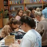 Osoby przy stole w bibliotece wykonują prace plastyczne. Półki z kolorowymi książkami w tle. Wnętrze jest jasne i przytulne.