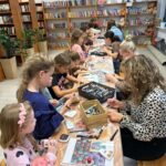 Grupa dzieci i dorosłych maluje kamienie przy drewnianym stole w bibliotece. Półki pełne książek i pluszaków w tle. Jasne wnętrze z roślinami.
