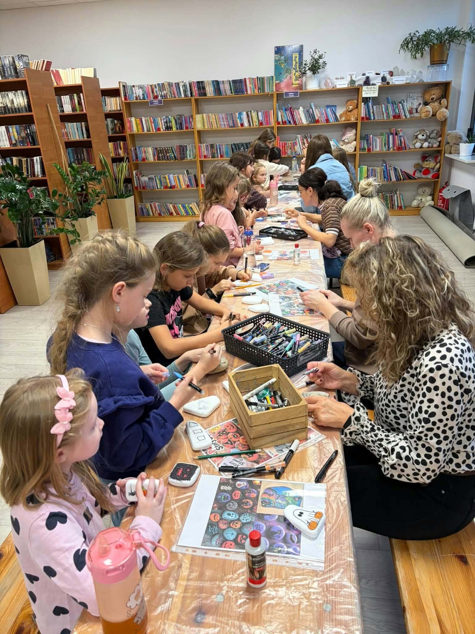 Grupa dzieci i dorosłych maluje kamienie przy drewnianym stole w bibliotece. Półki pełne książek i pluszaków w tle. Jasne wnętrze z roślinami.
